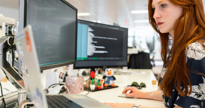 A professional woman wearing glasses sits at a desk working on three computers