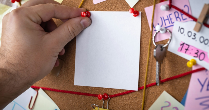 A hand pinning a blank piece of paper on a bulletin board with various notes and strings.